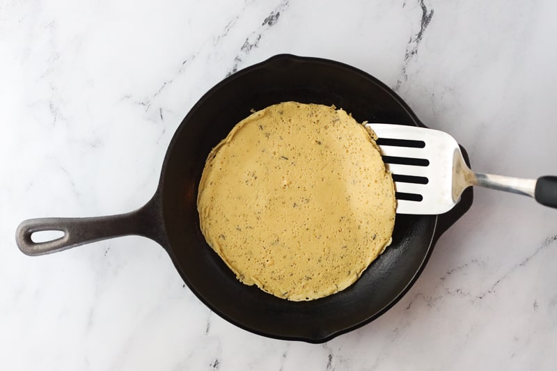Gluten-free chickpea flatbread batter in cast iron pan with spatula for flipping.