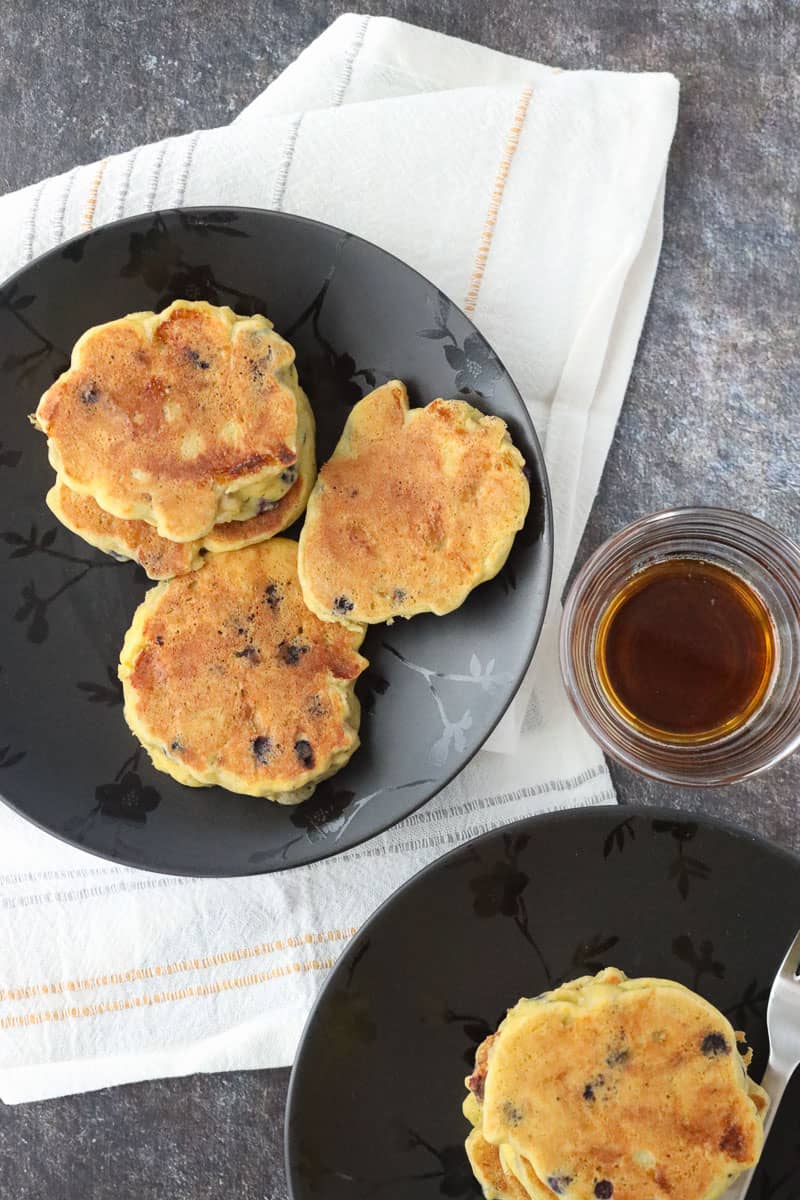 Cooked gluten-free corn flour pancakes on black floral plates with side of maple syrup.