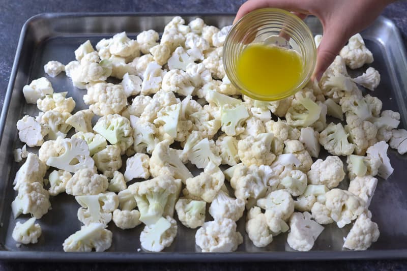 Cauliflower florets on a sheet pan with hand pouring butter from glass prep container.