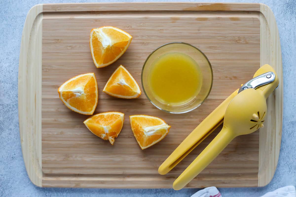 Halved orange wedges on a wood cutting board with a lemon squeezer and a small container of squeezed juice.