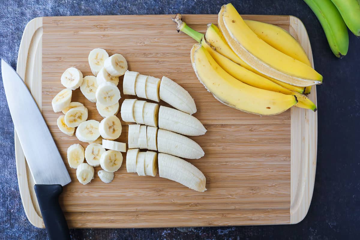 Partially sliced bananas on a cutting board with a chef's knife and banana peels on the side.