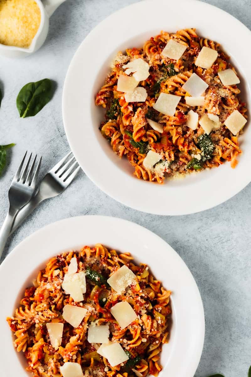 Fire-roasted tomato and artichoke pasta in two white bowls with shaved and grated cheeses sprinkled on top.