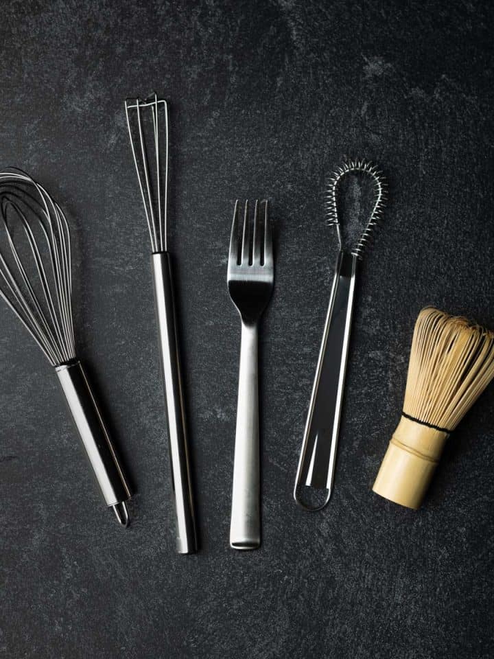 Ballon whisk, bar whisk, dinner fork, coil whisk, and bamboo whisk displayed on a black textured surface.