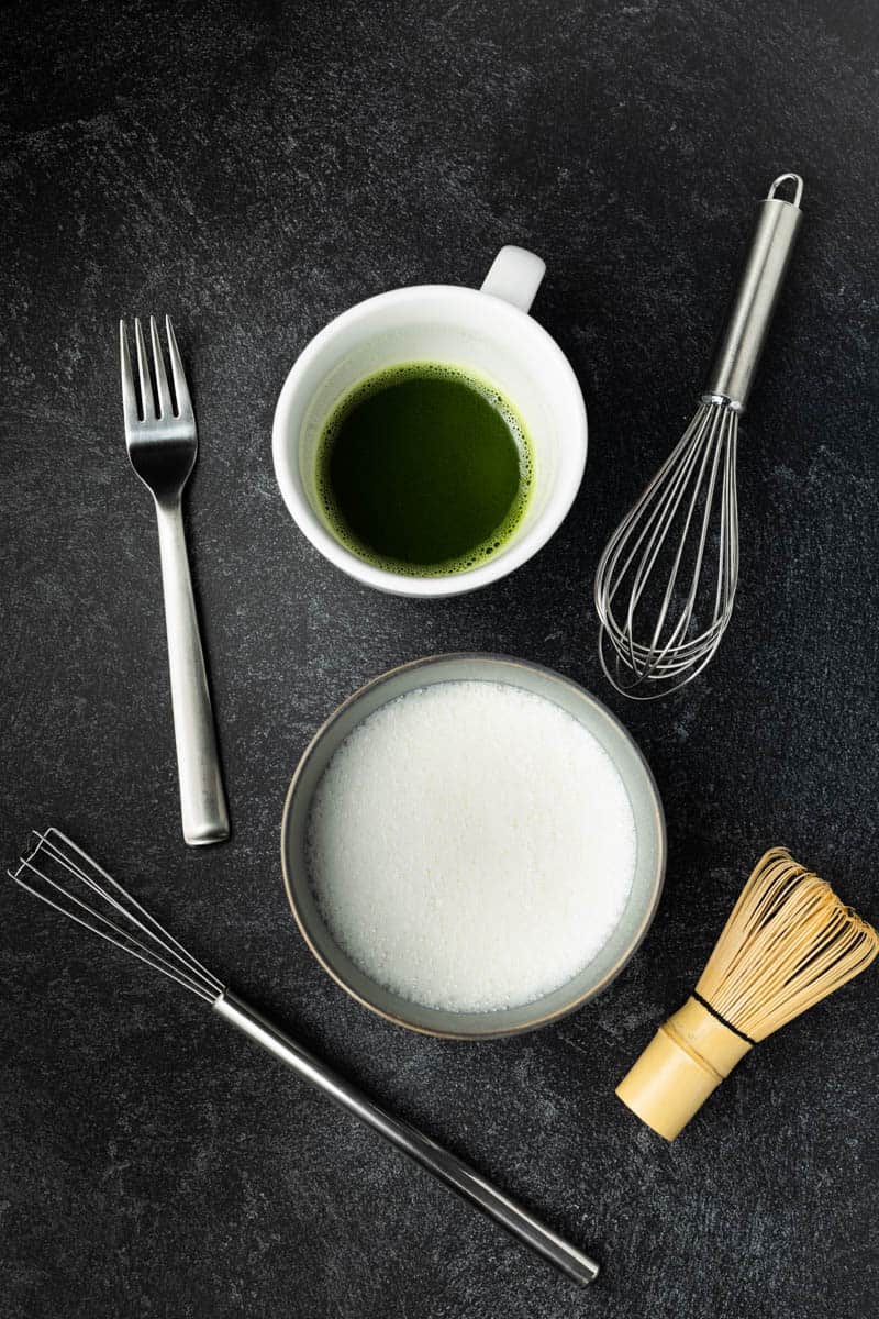 Frothed milk in a gray bowl next to matcha tea in a white cup, surrounded by various whisking tools.