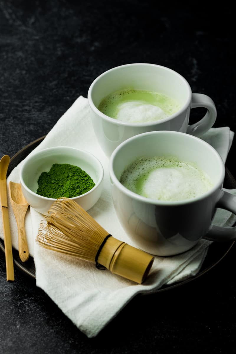 Two cups of vanilla matcha latte next to a small container of matcha powder, a bamboo whisk, a bamboo scoop, and a bamboo spoon.