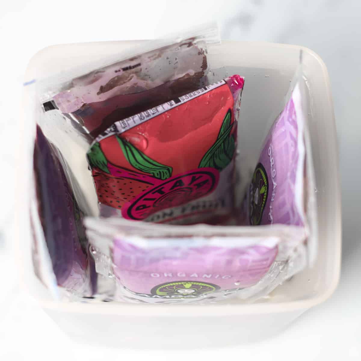 Packets of frozen acai puree and one packet of dragon fruit puree defrosting in a tall plastic container filled with hot water.