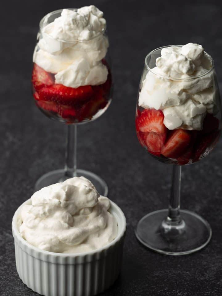 Two small wine glasses with chopped strawberries topped with homemade whipped cream, and one white ramekin full of whipped cream next to the glasses, on a textured charcoal background.