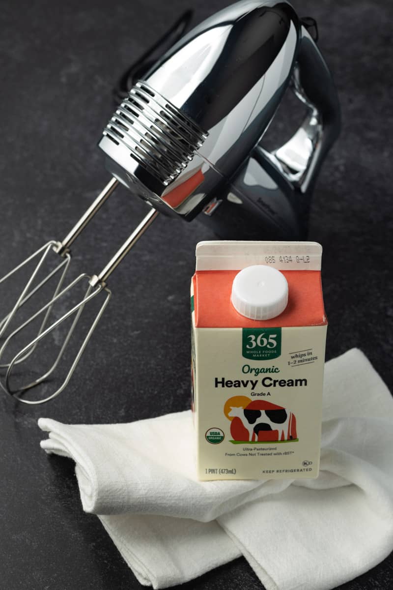 Retro-style handheld stainless-steel electric mixer next to a pint-sized carton of organic heavy cream resting on a white kitchen towel.