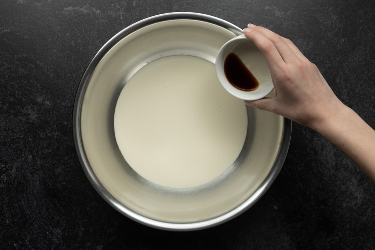 Adding vanilla extract to the heavy cream in a stainless-steel mixing bowl.