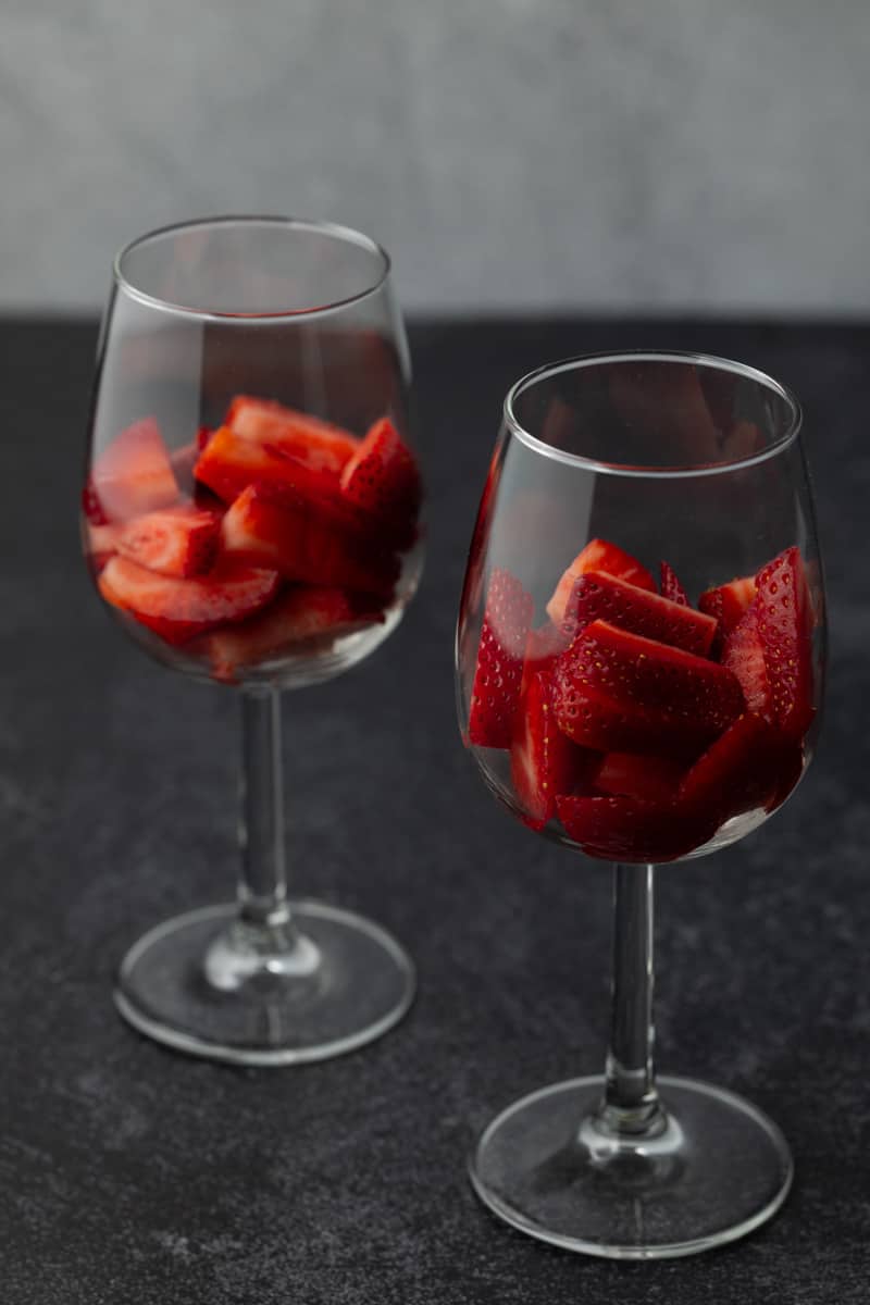 Two small wine glasses with fresh chopped strawberries on a textured charcoal surface and textured white background.