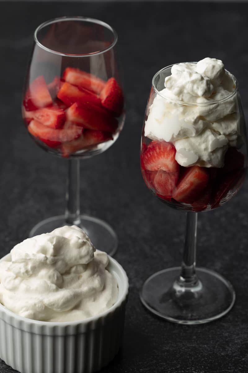 Two small wine glasses with chopped strawberries topped with homemade whipped cream, and one white ramekin full of whipped cream next to the glasses, on a textured charcoal background.