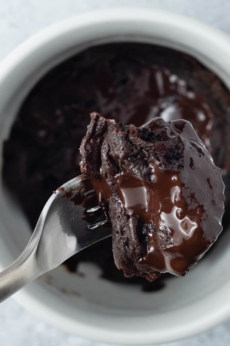 Top-down view of a chocolate protein powder mug cake in a white mug with a fork bite of cake in the foreground.