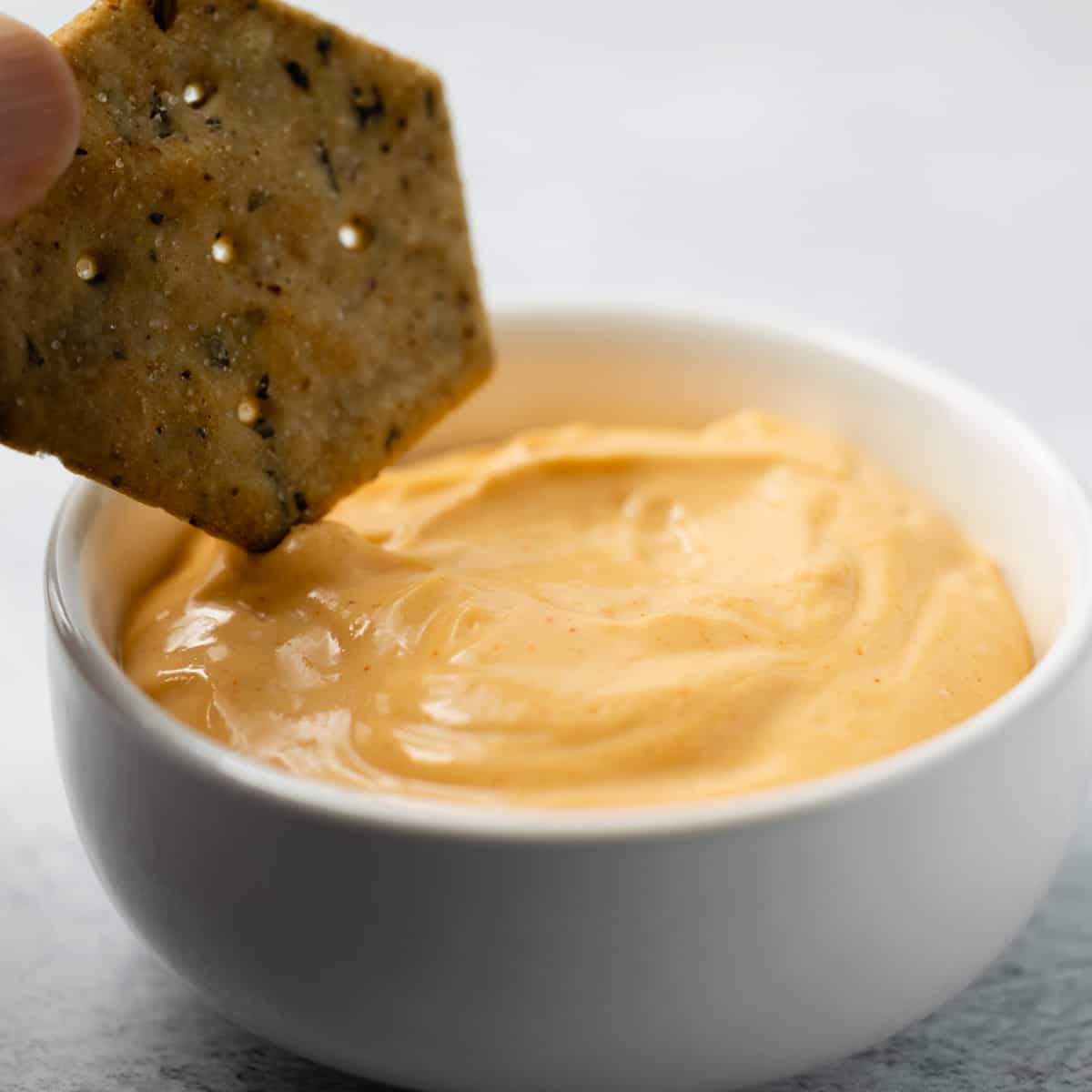 Flourless cheese sauce in a white bowl with a cracker dipping in.