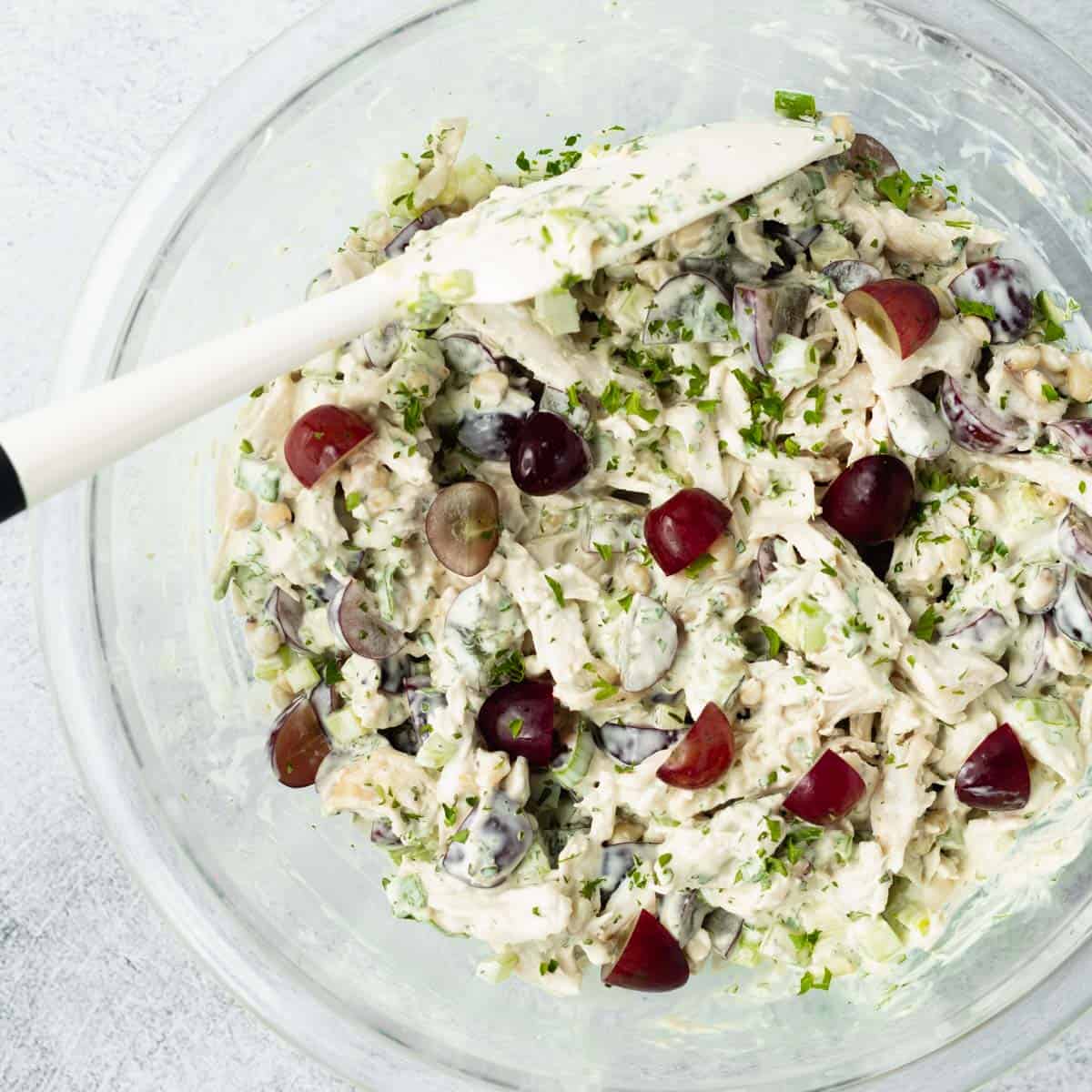 Finished chicken salad in a glass mixing bowl with a white spatula resting in the bowl.