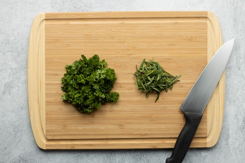 Bamboo cutting board with parsley and rosemary ready to be minced, next to a chef's knife on the right.