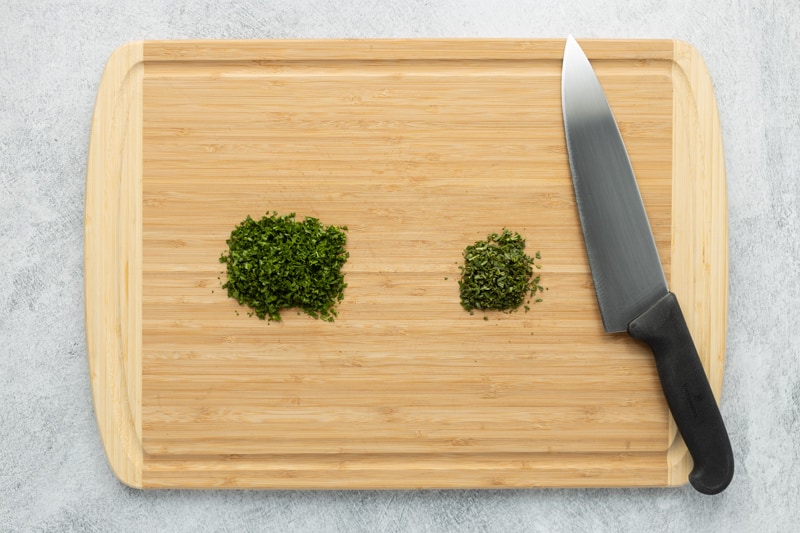 Bamboo cutting board with minced parsley and rosemary next to a chef's knife on the right.