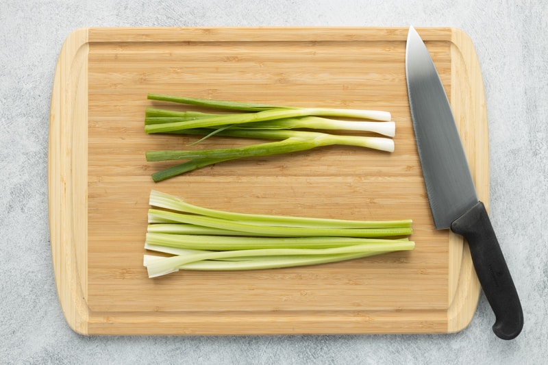 Bamboo cutting board with celery and green onions laid out ready to be cut with chef's knife on the right.
