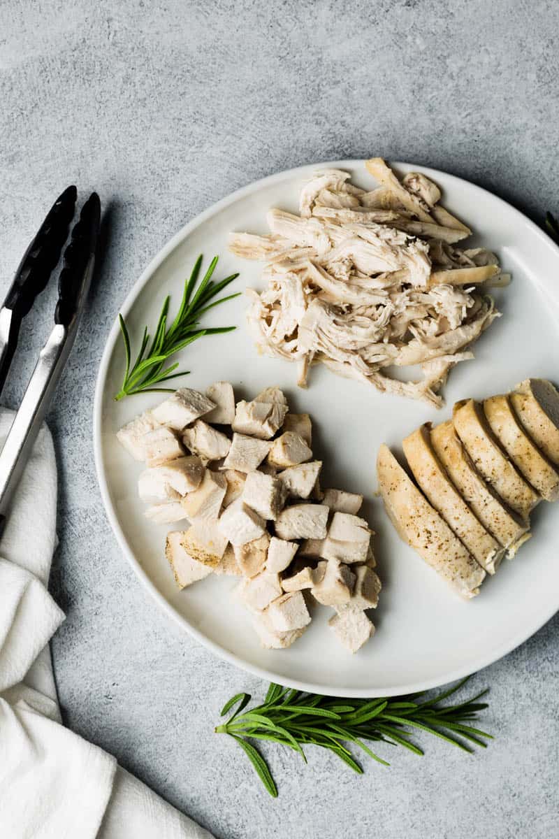 Cooked chicken breasts that have been cubed, shredded, and sliced, are placed on a white round platter, with snips of rosemary garnish and a pair of tongs on the side.