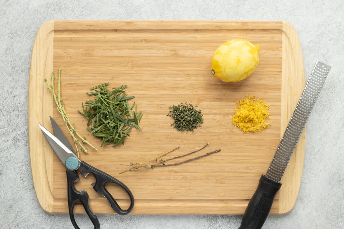 Destemmed rosemary and thyme and a zested lemon all resting on a cutting board next to a pair of kitchen scissors and a zester.