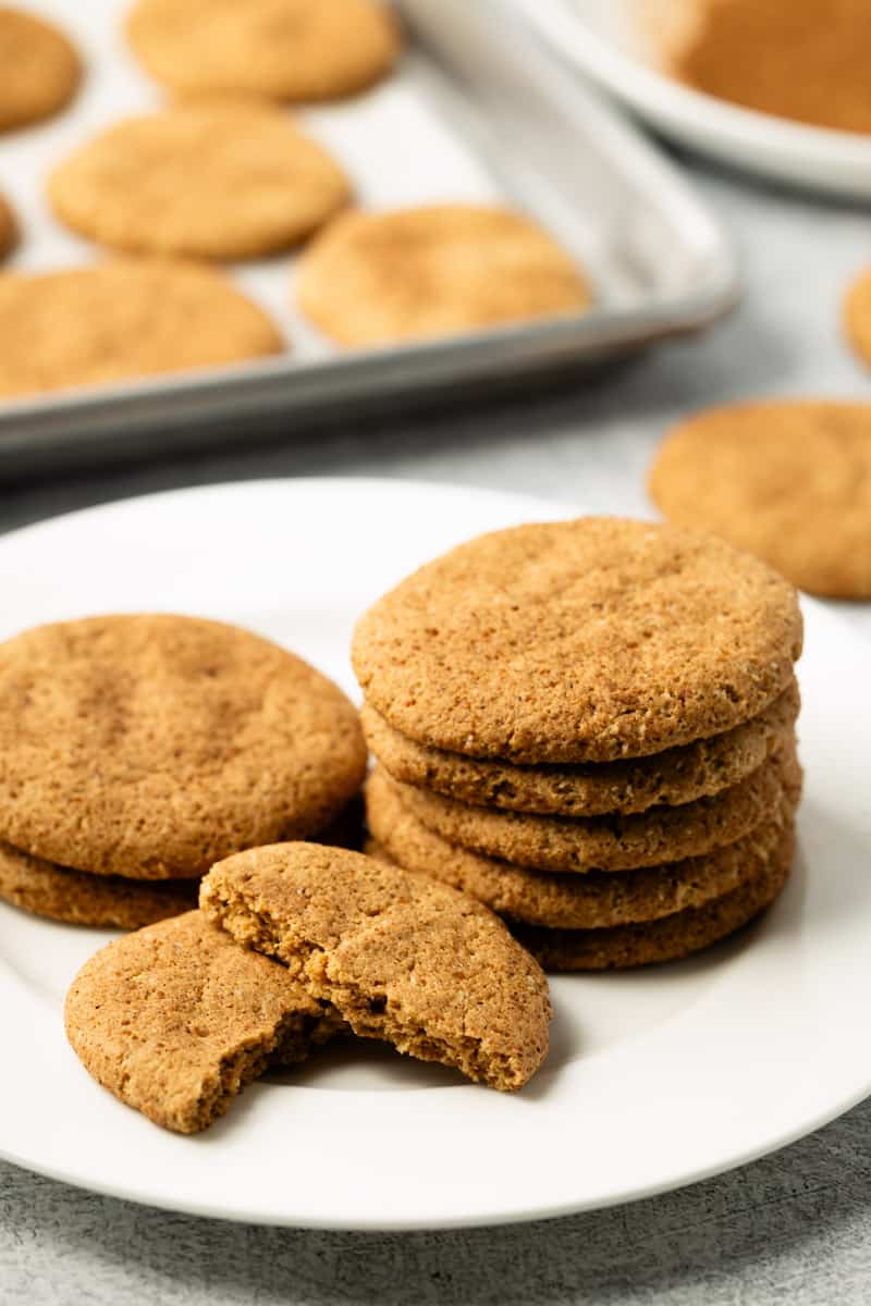 Protein snickerdoodle cookies stacked on a white plate with a baking sheet full of cookies in the background.