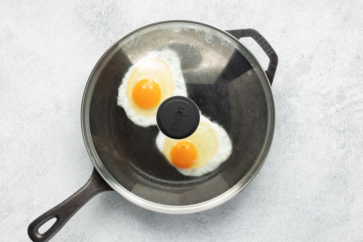 Two cracked eggs frying in a cast-iron skillet, covered with a glass lid.