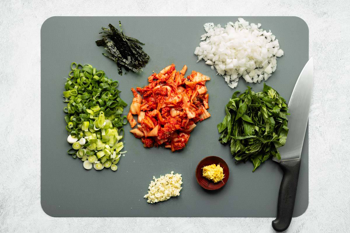 Chopped stir-fry veggies, sliced seaweed, ginger paste, and a chef's knife resting on a large gray cutting board.