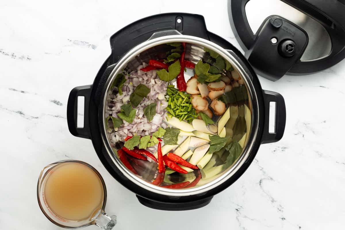 Chopped shallots, red chilies, herbs, and other aromatics are added to the Instant Pot with a measuring cup filled with chicken stock next to it.