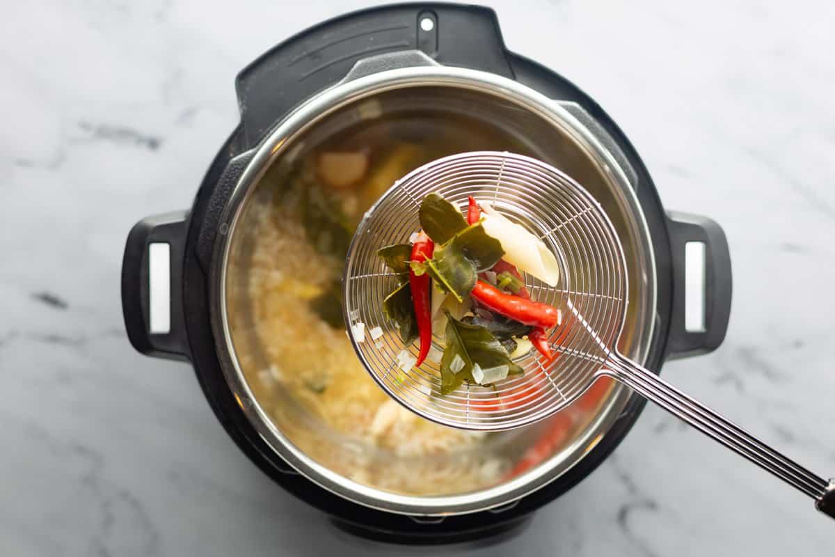 Instant Pot with the lid removed shows the pressure-cooked soup base ingredients with a strainer spoon over it containing the aromatics to be removed.
