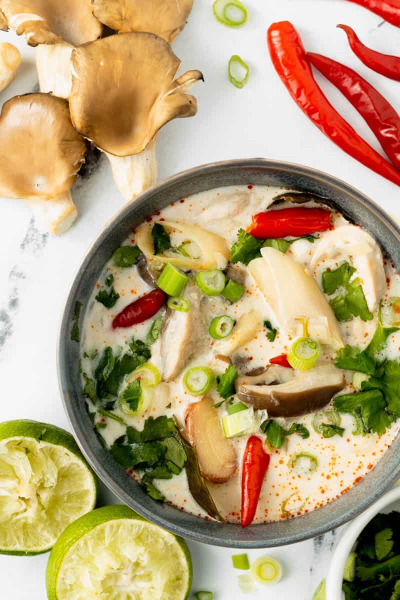 Thai Coconut Chicken Soup in a gray bowl is surrounded by mushrooms, lime, red chilies, green onions, and cilantro garnishes on a white marble background.