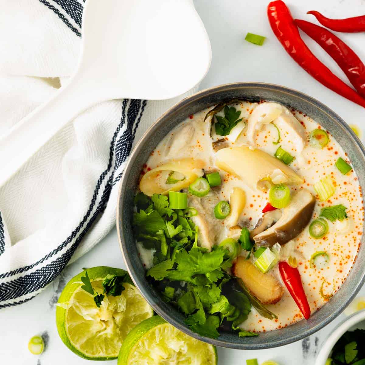 Finished Tom Ka Gai Soup in a gray bowl is surrounded by mushrooms, lime, red chilies, green onions, and cilantro garnishes on a white marble background.