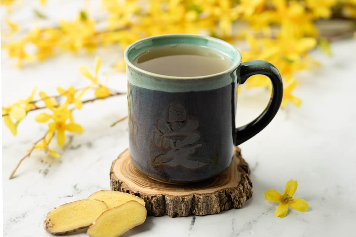 Lemongrass ginger tea in a ceramic mug with the Chinese character "love" on it, surrounded by yellow flowers and slices of ginger.