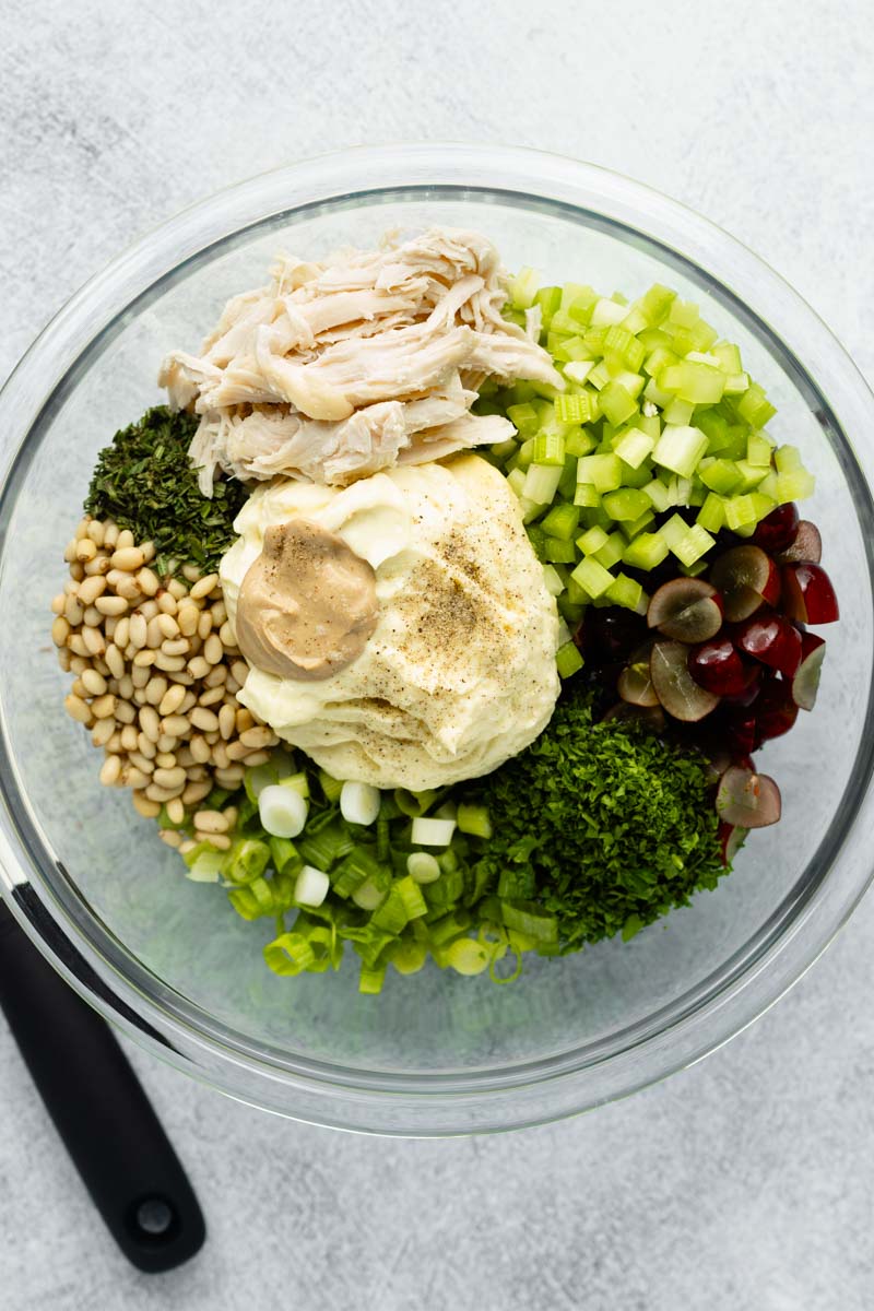 All gluten-free chicken salad recipe ingredients placed in a large glass mixing bowl, not yet mixed, with a white spatula resting on the left side.