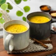 Two mugs of golden milk are resting on a light gray rope trivet with a walnut wood tray underneath.