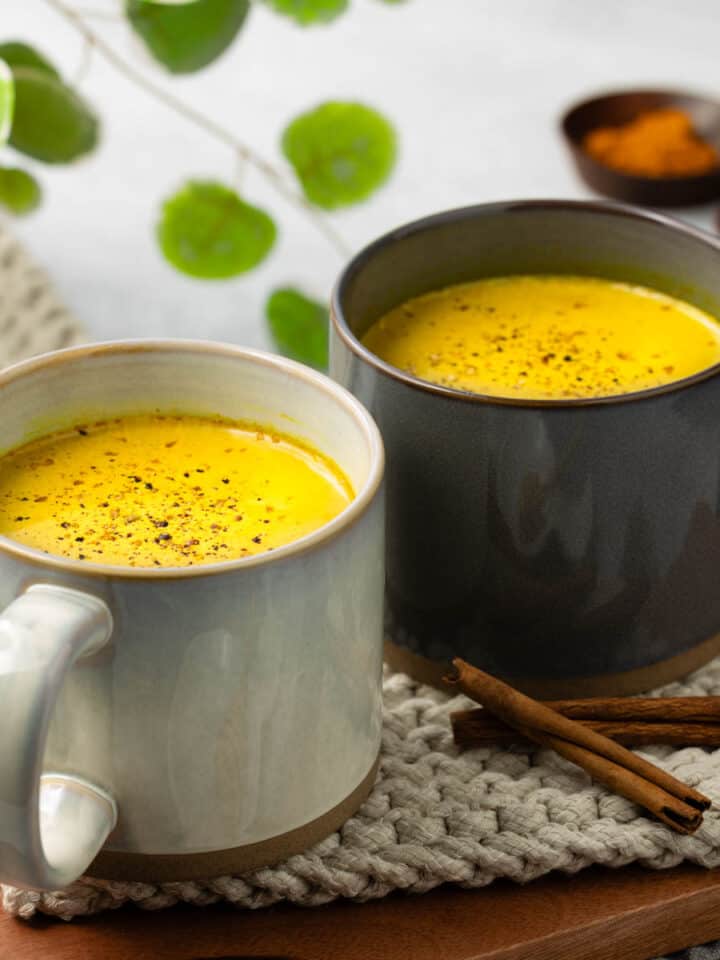 Two mugs of golden milk are resting on a light gray rope trivet with a walnut wood tray underneath.