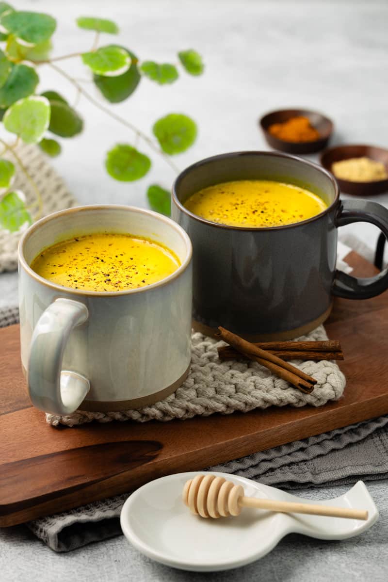 Two mugs of golden milk are resting on a light gray rope trivet with a walnut wood tray underneath.