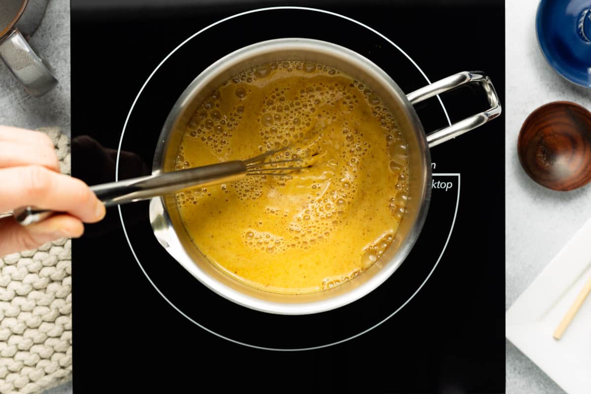 Golden milk ingredients are being whisked together in a small stainless-steel pot with a pour spout.