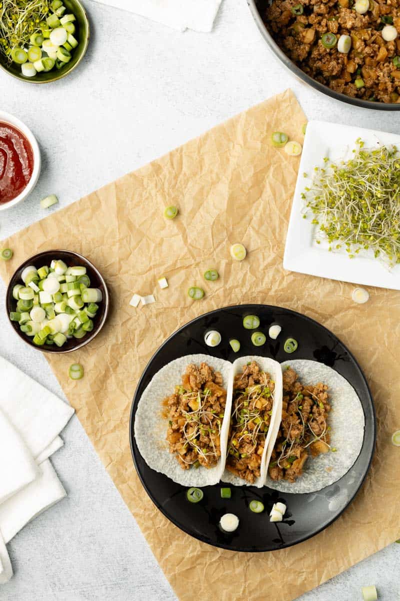 Three finished jicama tacos on a black plate are displayed next to extra ingredients on the side.