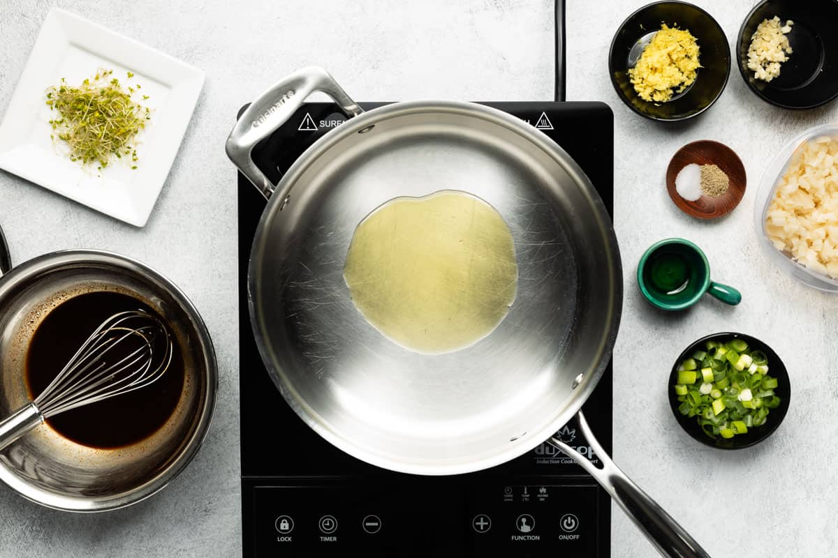 A stainless-steel skillet with avocado oil is heating up on a black cooktop with other recipe ingredients set to the side.