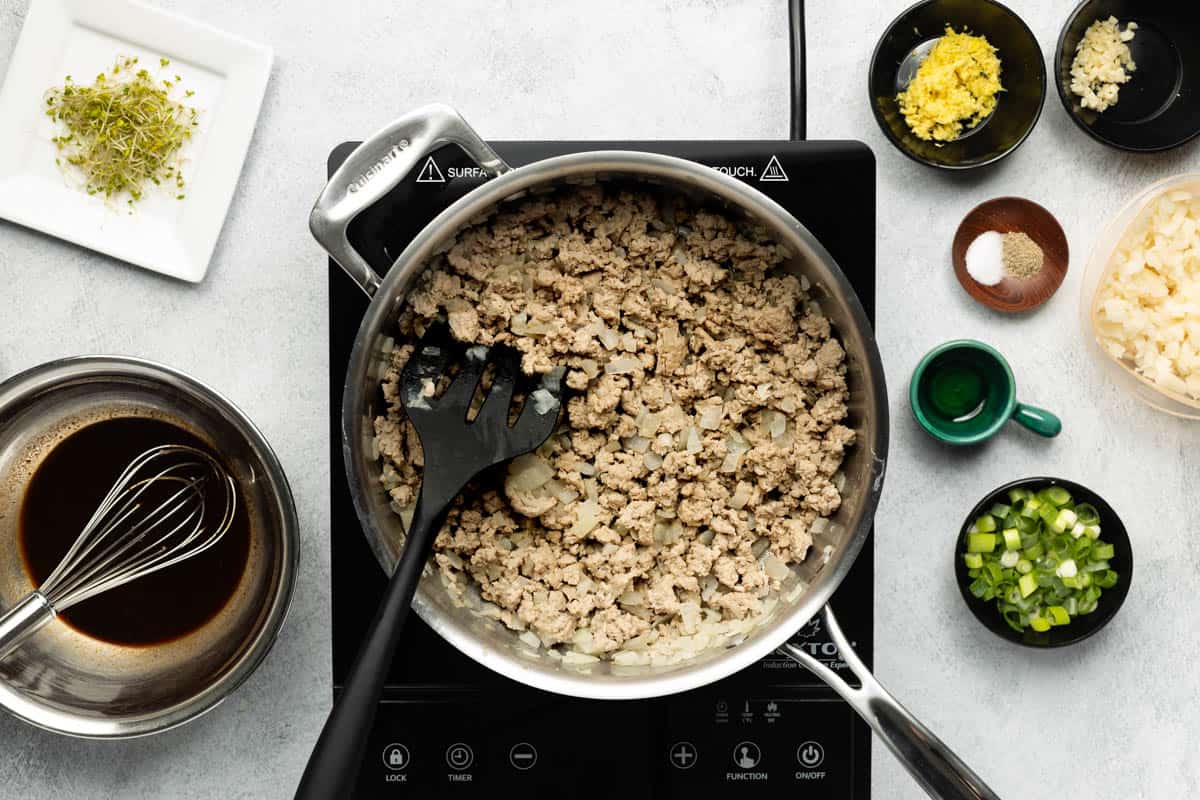 A stainless-steel skillet of cooked ground turkey added to the chopped onions is on a black cooktop with the remaining ingredients on the side.