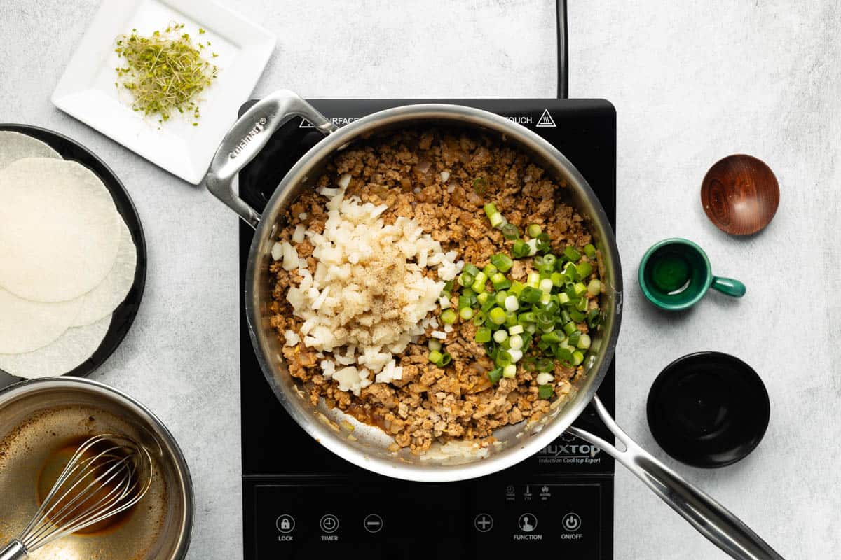 A stainless-steel skillet of cooked ground turkey with water chestnuts and green onions added on top is on a black cooktop with a plate of jicama wraps and broccoli sprouts on the side.