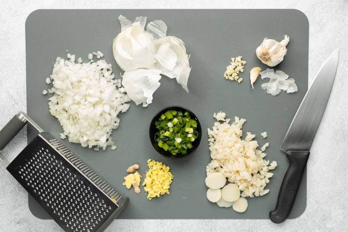 A large gray cutting board with chopped onions, garlic, water chestnuts, green onions, and grated ginger is resting on a gray textured surface with a chef's knife and grater on the side.