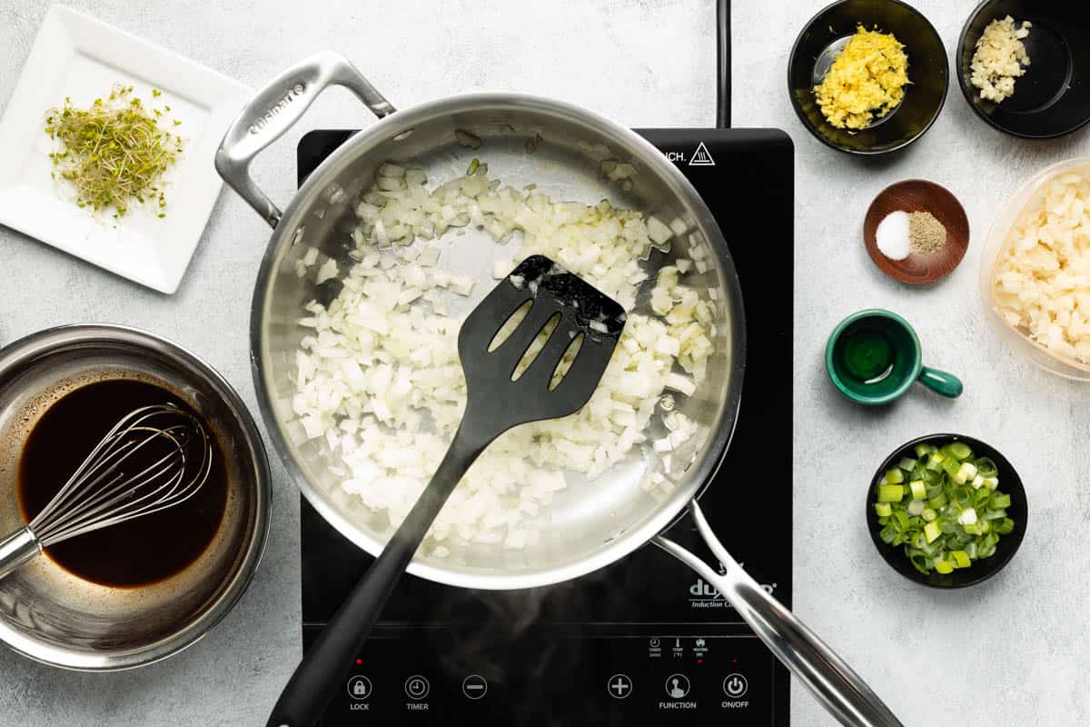 A stainless-steel skillet of chopped onions and avocado oil is cooking on a black cooktop with other recipe ingredients placed on the side.