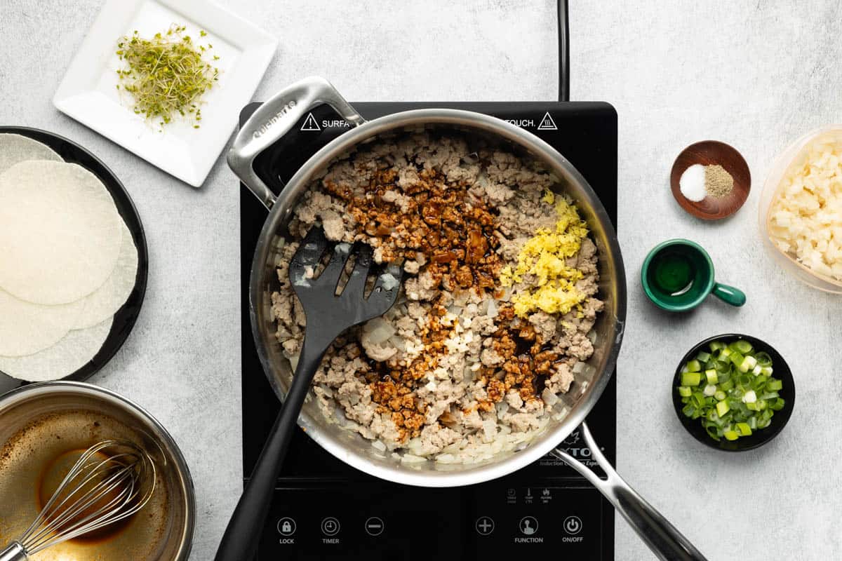 A stainless-steel skillet of cooked ground turkey combined with sauce and grated ginger is on a black cooktop with a plate of jicama wraps and remaining ingredients on the side.