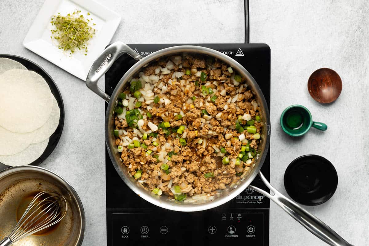 A stainless-steel skillet of cooked ground turkey combined with remaining ingredients is on a black cooktop with a plate of jicama wraps and broccoli sprouts on the side.