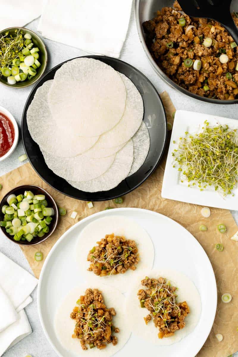 A spread of jicama wrap taco ingredients are above a white plate of jicama wraps with ground turkey filling.