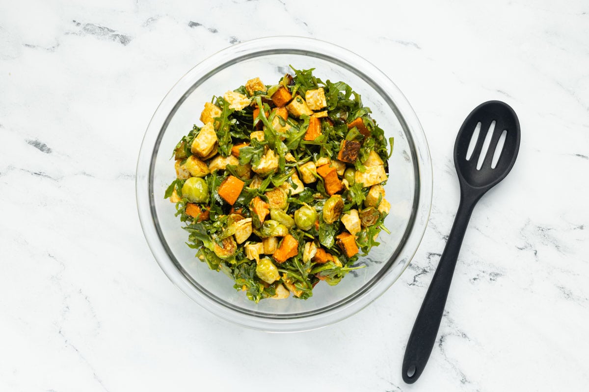 Curry chicken salad with roasted vegetables combined with curry vinaigrette in a glass mixing bowl, next to a large black slotted spoon.