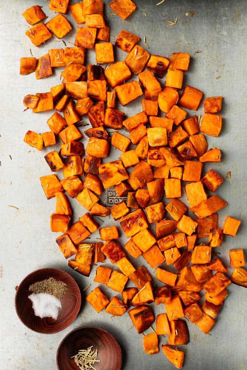 Crispy oven-roasted sweet potatoes are on a baking sheet next to two small wooden prep bowls with rosemary, salt, and pepper.