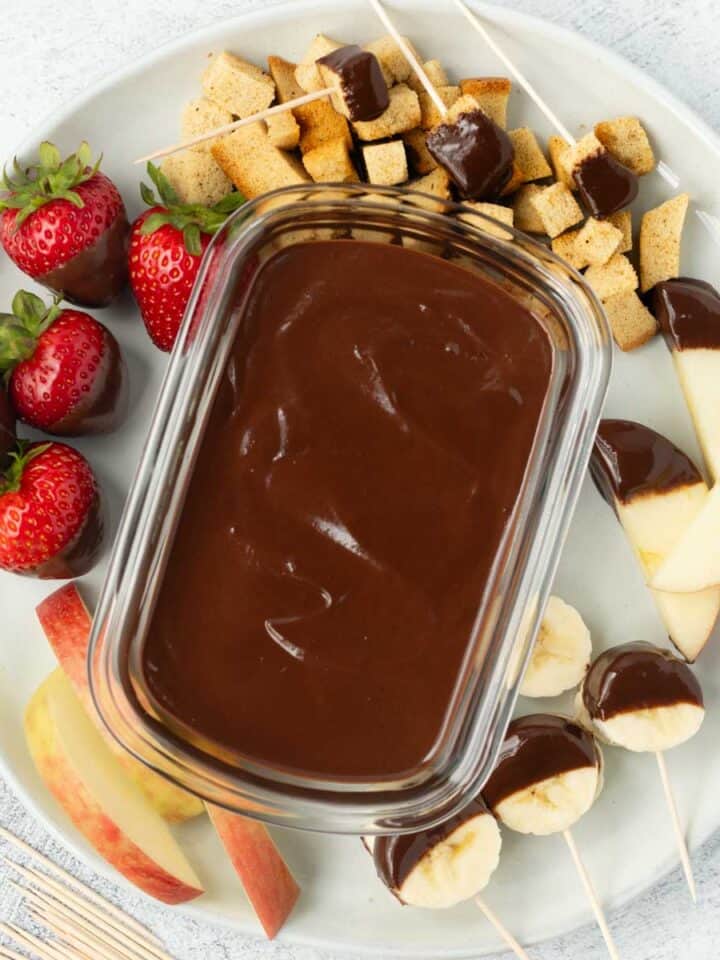 A container of homemade chocolate sauce on a plate surrounded by fruit and bread cubes.