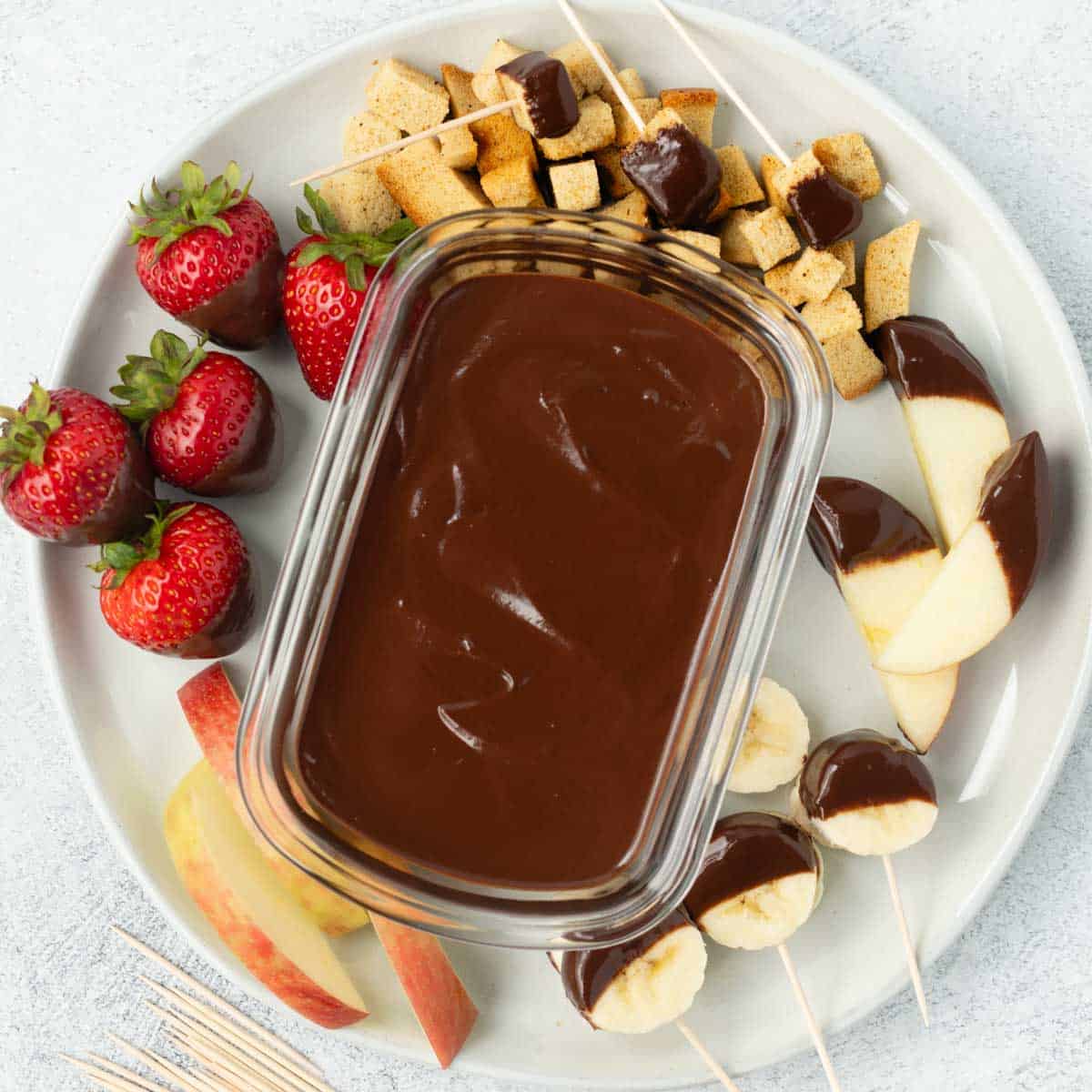 A container of homemade chocolate sauce on a plate surrounded by fruit and bread cubes.