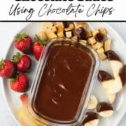 A container of homemade chocolate sauce on a plate surrounded by fruit and bread cubes.